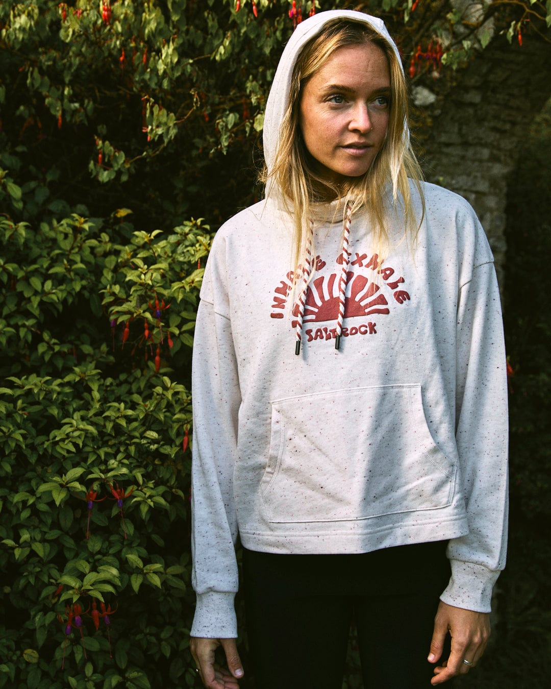 A person with long hair stands outdoors, wearing the Kellie - Women's Cropped Pop Hoodie in cream by Saltrock and dark pants. Green foliage and flowers bloom in the background.