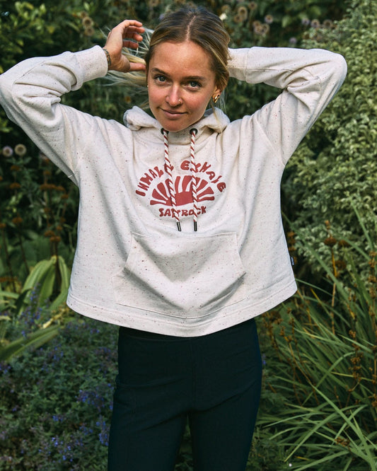 A person in the Kellie Women's Cropped Pop Hoodie in cream from Saltrock stands outdoors, holding their hair up with both hands.