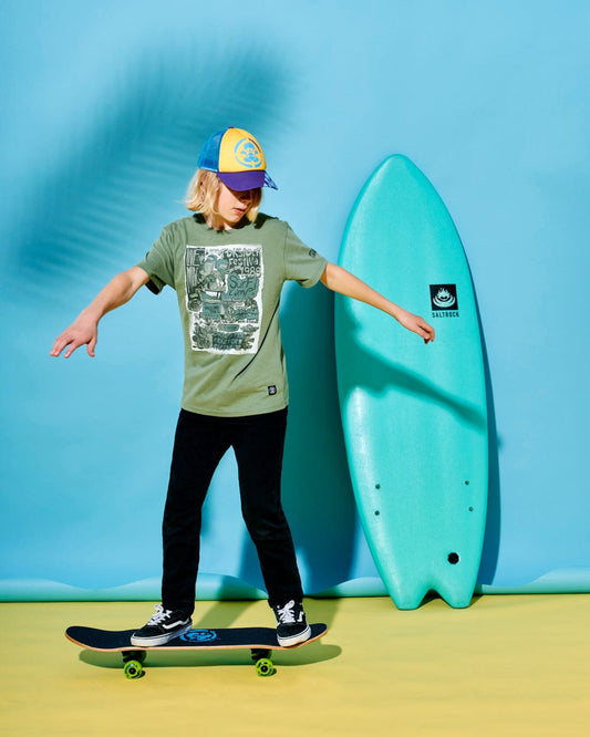 A child with long blonde hair wearing a Saltrock Festival Merch - Kids Short Sleeve T-Shirt - Green, black pants, and a colorful baseball cap balances on a skateboard in front of a blue backdrop with a turquoise surfboard. The T-shirt is made from 100% cotton for ultimate comfort.