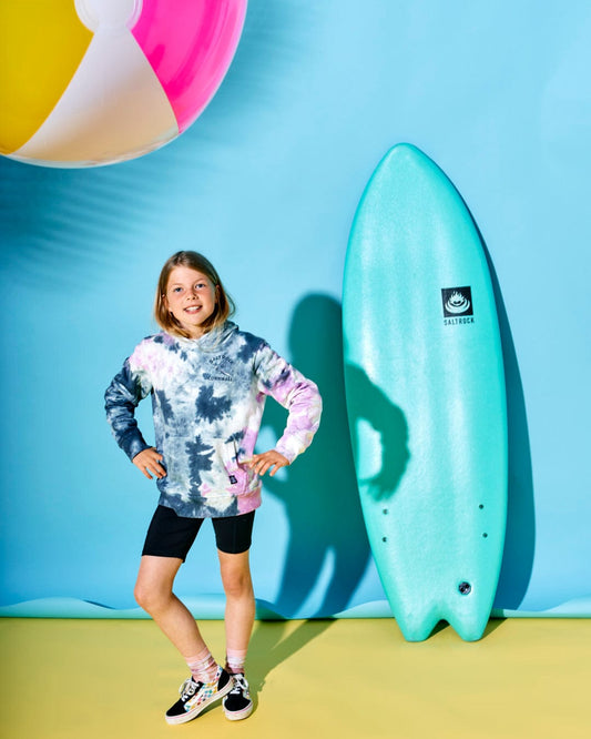 A child stands in front of a surfboard wearing a Wave Rider Cornwall - Kids Tie Dye Pop Hoodie - Pink/grey and shorts. There is a large beach ball with the Saltrock Running Man logo and a blue background.