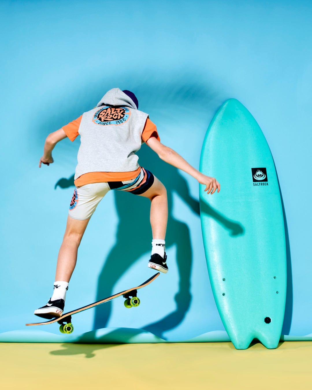 A person in a Saltrock SR Original - Kids Sleeveless Pop Hoodie - Grey and shorts is mid-air on a skateboard. A turquoise surfboard stands upright against a bright blue background.