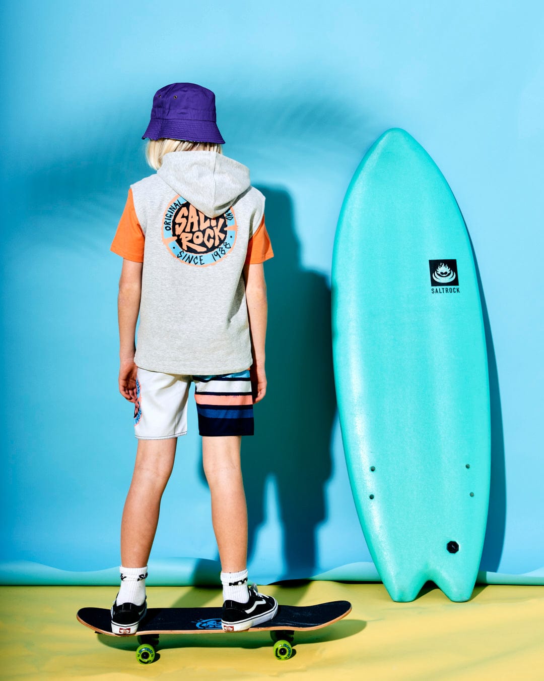 Child with blonde hair wearing a Saltrock SR Original - Kids Sleeveless Pop Hoodie - Grey and shorts stands on a skateboard facing away. A turquoise surfboard is leaned against a blue background, adding a touch of coastal charm reminiscent of Saltrock graphics.