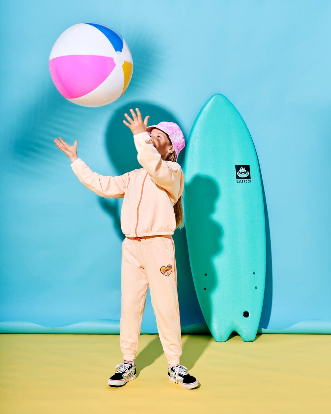 A person in pastel sportswear and a pink cap tosses a beach ball. Clad in Saltrock branded Sunshine State - Kids Joggers - Peach with an elasticated waistband, they stand before a surfboard leaning against a blue wall behind them.