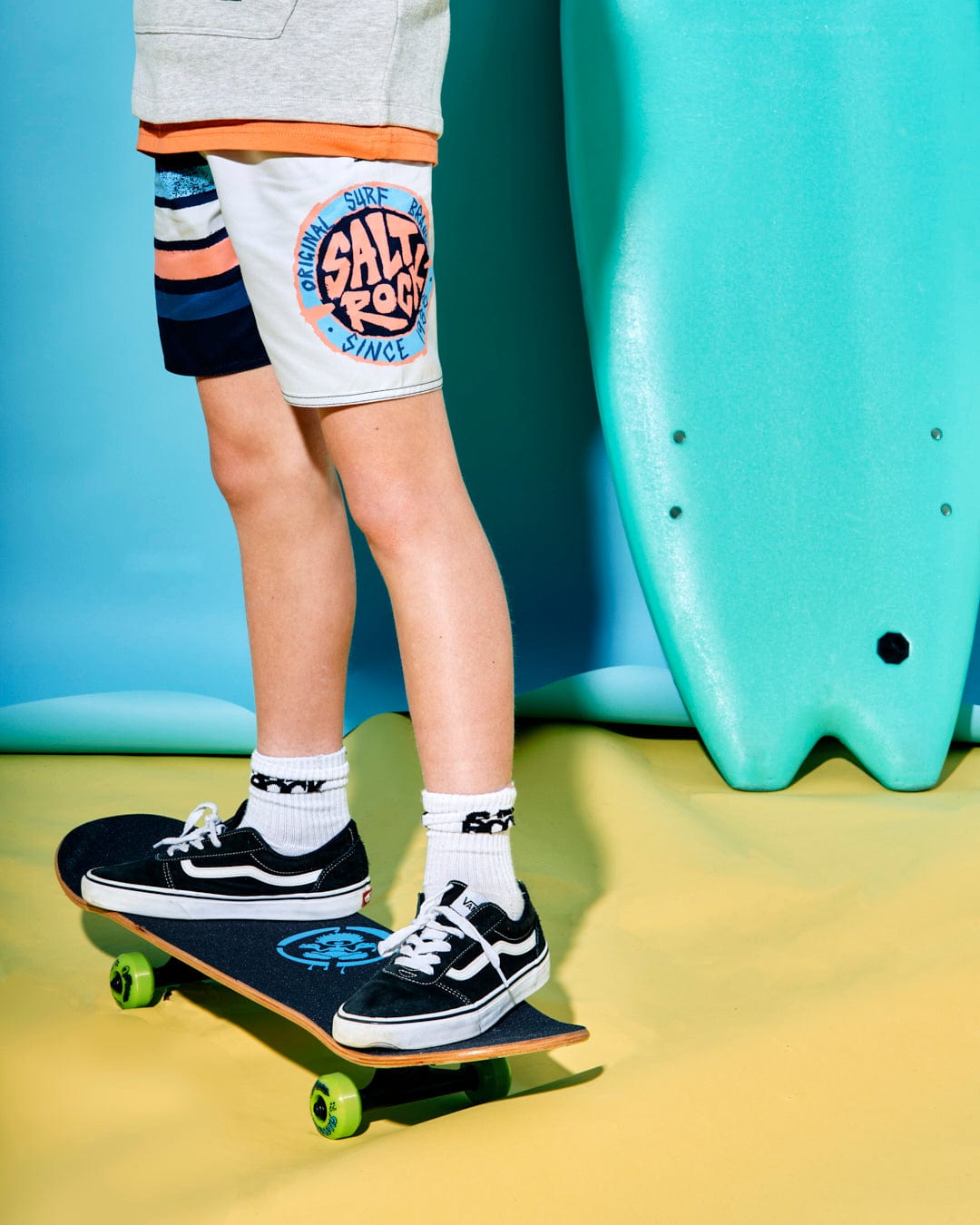 Young person wearing Saltrock Original SR - Kids Swimshorts - Blue with an elasticated waist and black sneakers, standing on a skateboard next to a bright turquoise surfboard, against a blue and yellow backdrop.