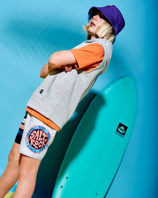 A person wearing a bucket hat, orange shirt, Saltrock SR Original - Kids Sleeveless Pop Hoodie - Grey, and colorful shorts stands with arms crossed next to a turquoise surfboard against a blue background.