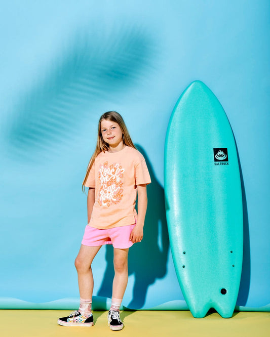 A child wearing a peach base T-shirt and pink shorts stands next to a turquoise surfboard against a light blue background with a shadow of a palm leaf. The vibrant outfit made of 100% cotton radiates the carefree spirit of Peace Love - Kids Short Sleeve T-Shirt - Peach by Saltrock.