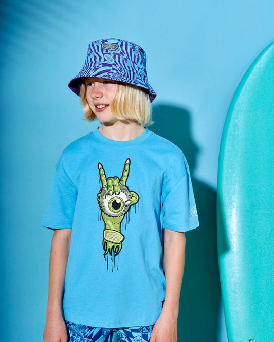 A person with long hair wearing a blue graphic t-shirt and a Warp Icon - Kids Bucket Hat - Blue by Saltrock featuring a geometric print stands next to a surfboard against a blue backdrop.