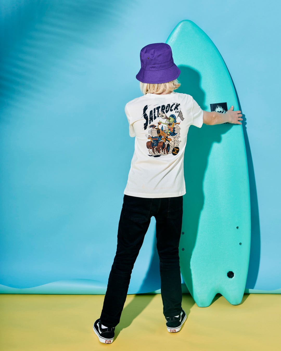 A person with blonde hair, wearing a purple bucket hat, black pants, and a white 100% cotton No Road No Problem - Kids Short Sleeve T-Shirt - White by Saltrock, stands facing a blue surfboard on a yellow and blue background.