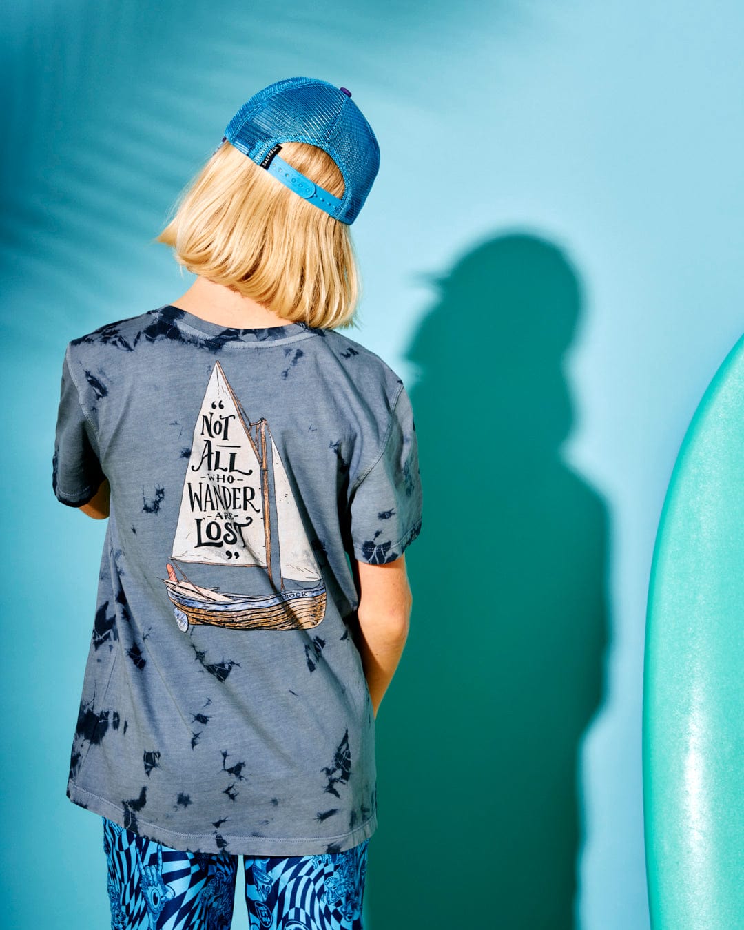 A child wearing a blue tie-dye Lost Ships - Kids Short Sleeve T-Shirt from Saltrock with a sailboat graphic and the text "Not All Who Wander Are Lost" on the back. The child is also wearing a blue cap and stands beside a turquoise surfboard against a blue background.