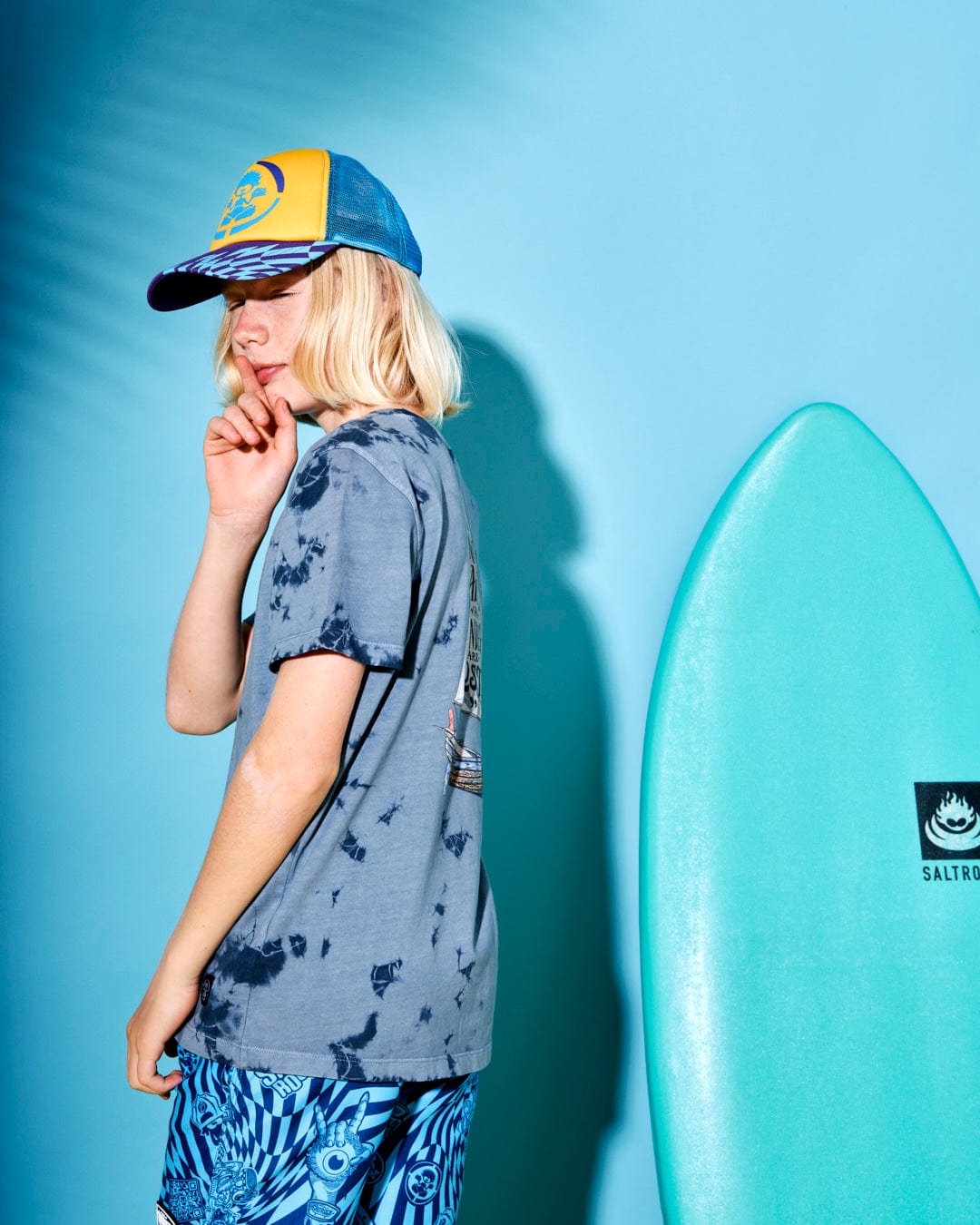 A person wearing a cap and casual beachwear stands next to a light blue surfboard with a hint of blue tie dye against a solid blue background, showcasing the Saltrock Lost Ships - Kids Short Sleeve T-Shirt - Blue Tie Dye.