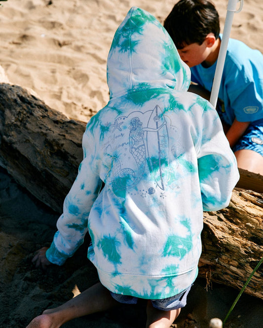 Child in a white and teal tie-dye hoodie with a shimmering silver foil mermaid print on the back, sitting on the beach near a log with a companion nearby, wearing the Mermaid Surf - Kids Tie Dye Pop Hoodie - Turquoise/White by Saltrock.