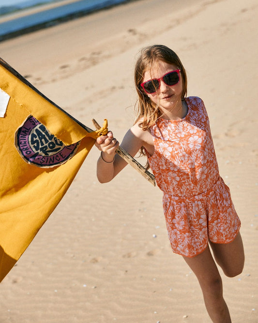A young girl wearing pink sunglasses and a loose fit **Saltrock Hibiscus - Kids Playsuit - Pink/Orange** holds a yellow flag on a sandy beach.