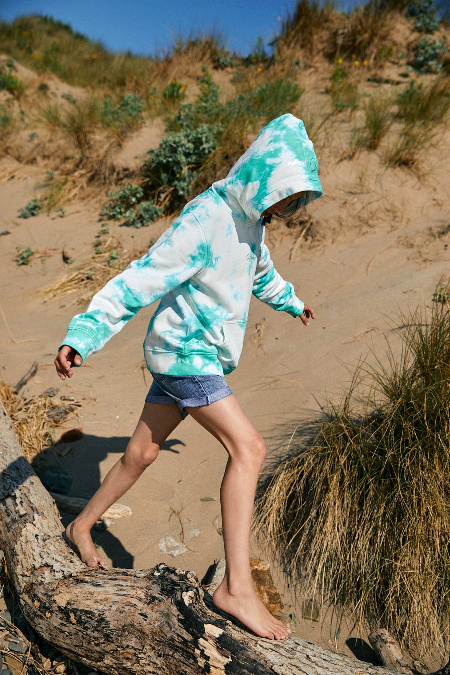A person wearing a Saltrock Mermaid Surf - Kids Tie Dye Pop Hoodie - Turquoise/White and shorts walks barefoot on a sandy terrain with sparse vegetation and a fallen tree branch.
