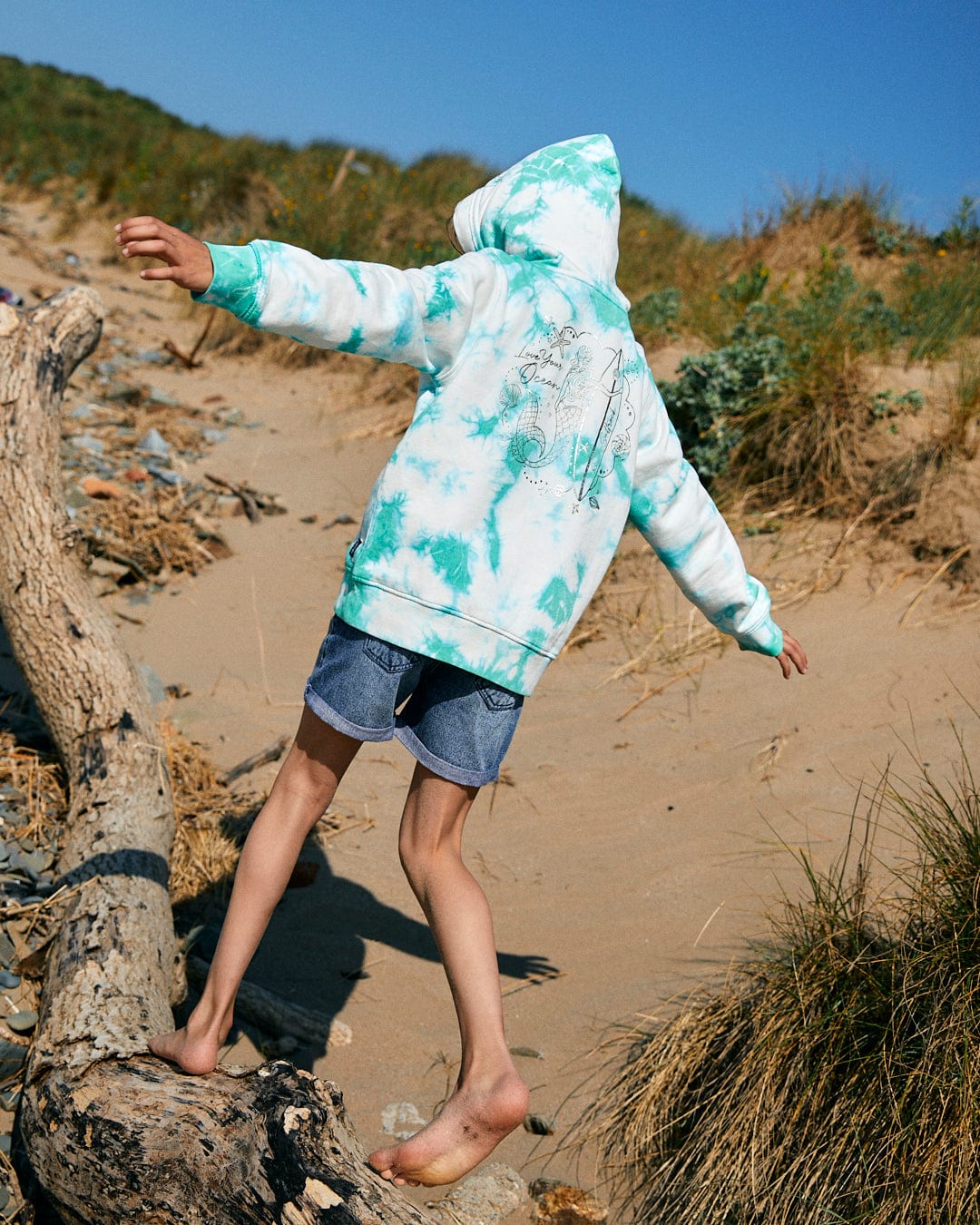 Person in a Saltrock Mermaid Surf - Kids Tie Dye Pop Hoodie - Turquoise/White and denim shorts balances barefoot on a log on a sandy beach.