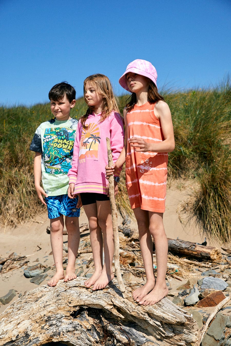 Three children, dressed in Saltrock Retro Seascape - Recycled Kids Longline Pop Hoodie - Pink, stand barefoot on a large piece of driftwood at a beach, with grass-covered dunes in the background. One child holds a stick while their vibrant outfits feature playful palm prints.