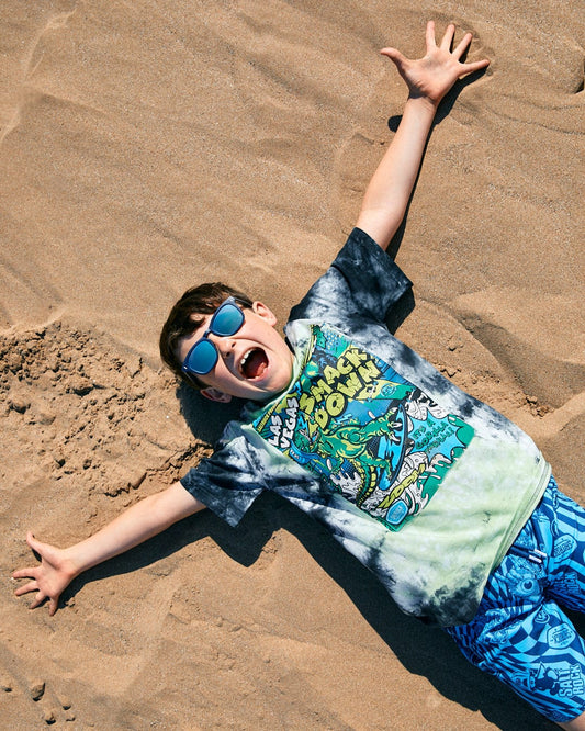 A child wearing sunglasses and a Las Vegas Smackdown - Kids Glow in the Dark T-Shirt - Multi by Saltrock is lying on the sand with arms and legs stretched out, appearing to yell or express excitement.