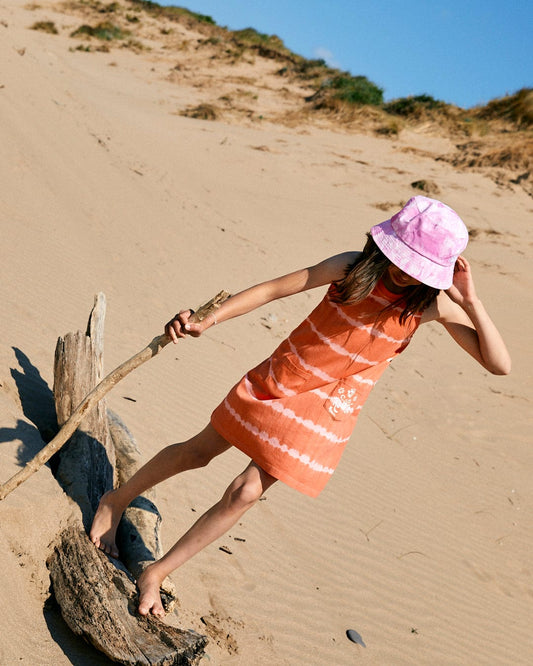 A child in a sleeveless, Eliana - Kids Tie Dye Vest Dress - Peach by Saltrock balances on a driftwood log on a sandy beach while holding a stick, wearing a pink bucket hat.