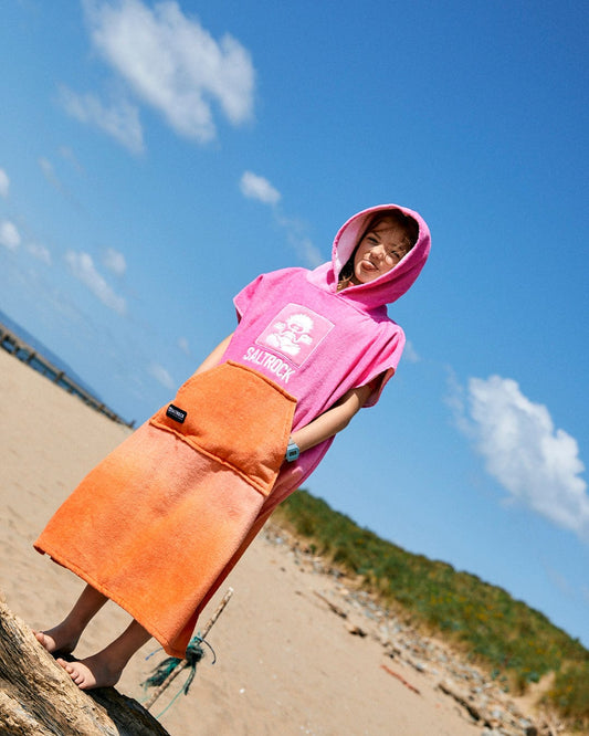 A child wearing a Saltrock Tropic Dip - Kids Changing Towel - Pink/Orange stands on a sandy beach with one foot on a log. The ultra-absorbent fabric keeps them dry as their eyes close in contentment under the clear sky with a few clouds.