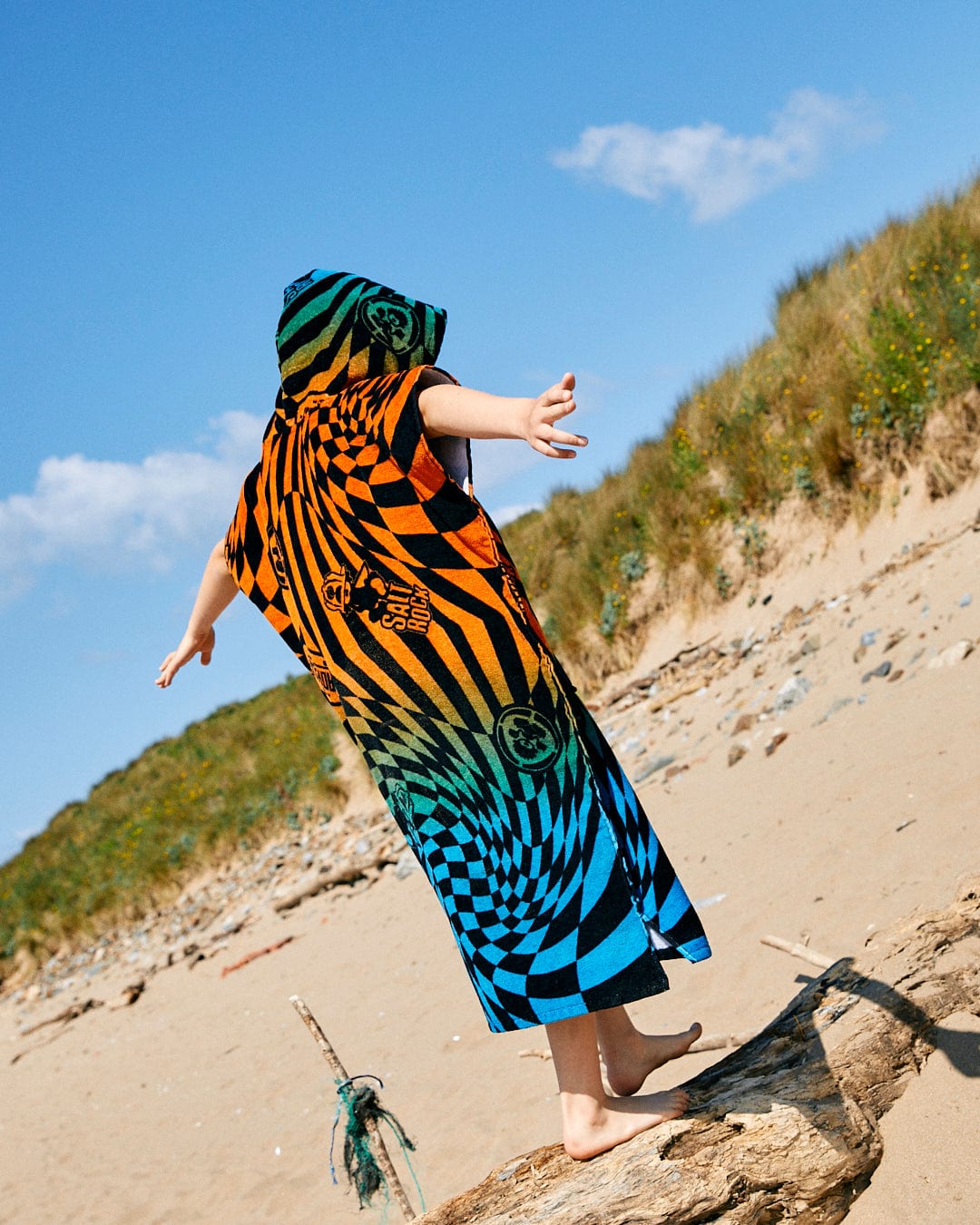 A child stands on a log at the beach, wearing an oversized Saltrock Warp Icon - Kids Changing Towel - Blue/Orange with arms outstretched. Sandy dunes and vegetation frame the background under a blue sky, while the towel's colorful pattern complements its fast-drying towelling material.