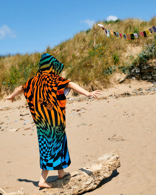 A person in a Saltrock Warp Icon - Kids Changing Towel - Blue/Orange balances on a log at a sandy beach, with grassy dunes and prayer flags visible in the background.