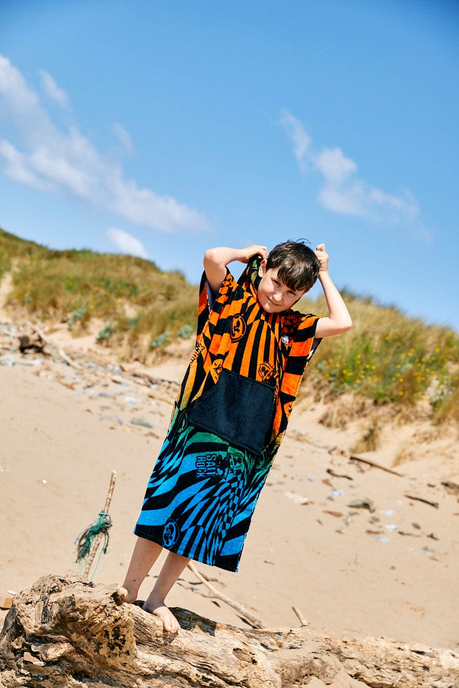 A child stands on a sandy beach, wrapped in an oversized Saltrock Warp Icon - Kids Changing Towel - Blue/Orange with colorful patterns over their clothes. The background displays dunes and greenery under a clear blue sky.