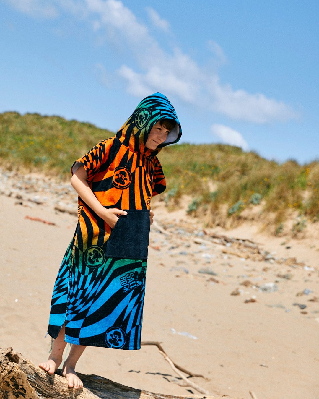 A child stands on a sandy beach wearing a Saltrock Warp Icon - Kids Changing Towel - Blue/Orange with patterns, against a backdrop of grassy dunes and a clear blue sky.