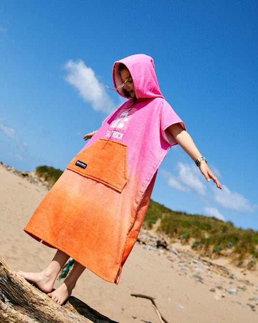 A child wearing a Saltrock Tropic Dip - Kids Changing Towel - Pink/Orange stands on a sandy beach, balancing on a log under a clear blue sky.