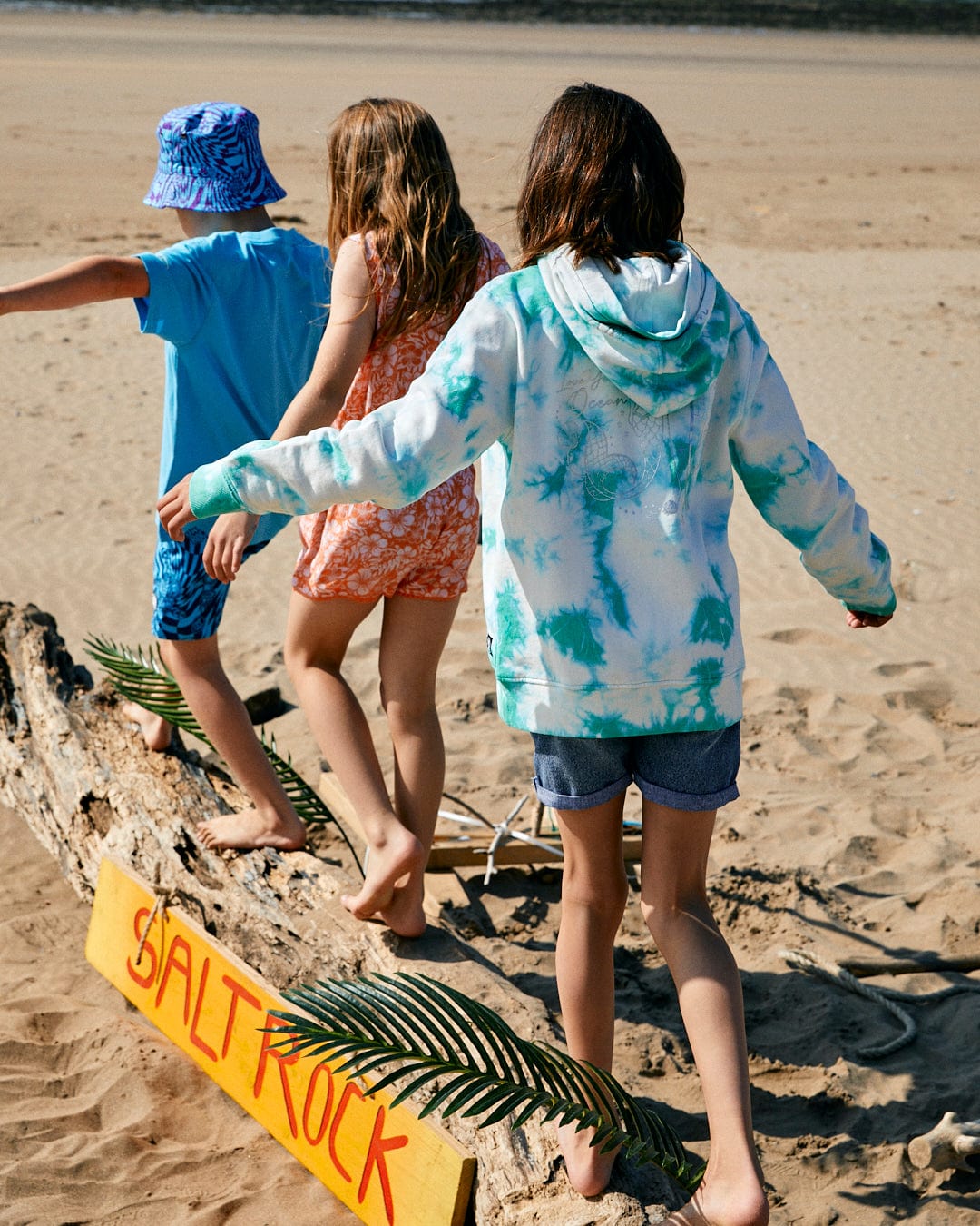 Three children balance on a large piece of driftwood on a sandy beach. A yellow sign with "SALTROCK" written in red underscores the Saltrock branding, while hints of tie dye patterns from the Mermaid Surf - Kids Tie Dye Pop Hoodie - Turquoise/White by Saltrock dance in the background.
