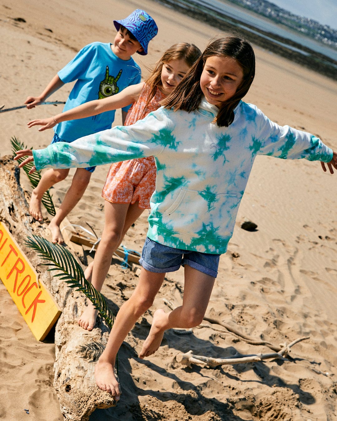 Three children balancing on a driftwood log on the beach. One wears a Mermaid Surf - Kids Tie Dye Pop Hoodie - Turquoise/White with Saltrock branding, another an orange dress, and the third a blue T-shirt and hat. A yellow sign partially visible reads "ROCK.