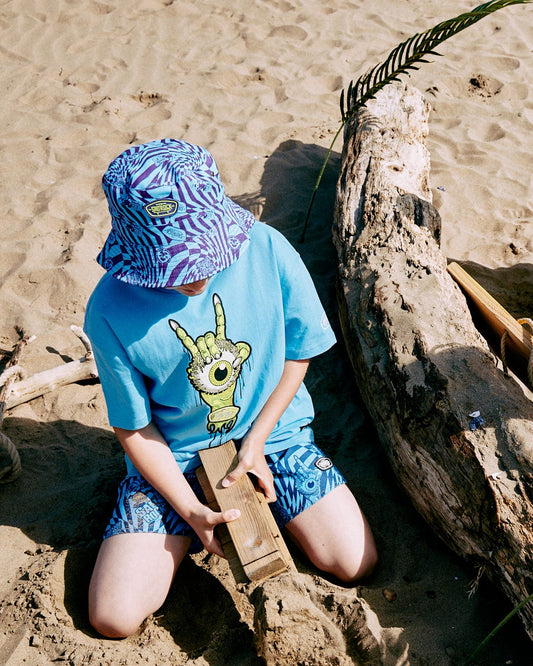 A child wearing a blue, 100% cotton Bowling for Surf - Kids Short Sleeve Glow in the Dark T-Shirt by Saltrock and a patterned hat digs in the sand with a wooden block next to a large piece of driftwood on a sandy beach.