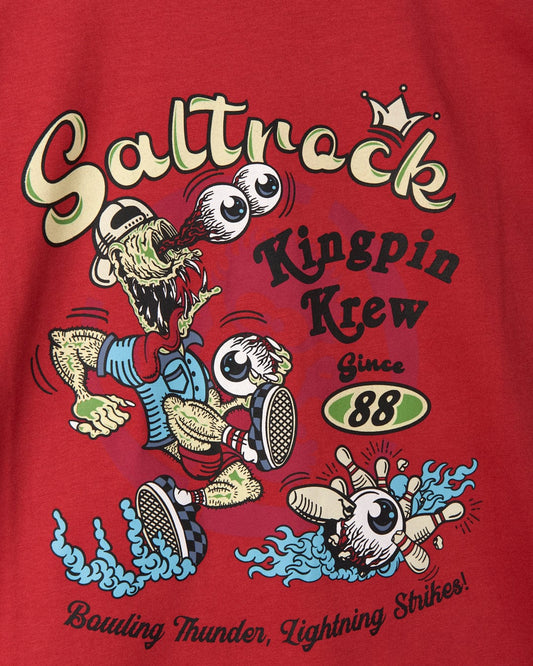 A colorful, vintage-style graphic on a red shirt features the words "Saltrock Kingpin Krew Since '88" with a cartoon character being chased by a surfboard and waves. This relaxed fit tee also has the added fun of glow-in-the-dark details, accompanied by "Boulting Thunder, Lightning Strikes! The Kingpin Krew - Kids Short Sleeve Glow in the Dark T-Shirt from Saltrock is perfect for playful adventures.