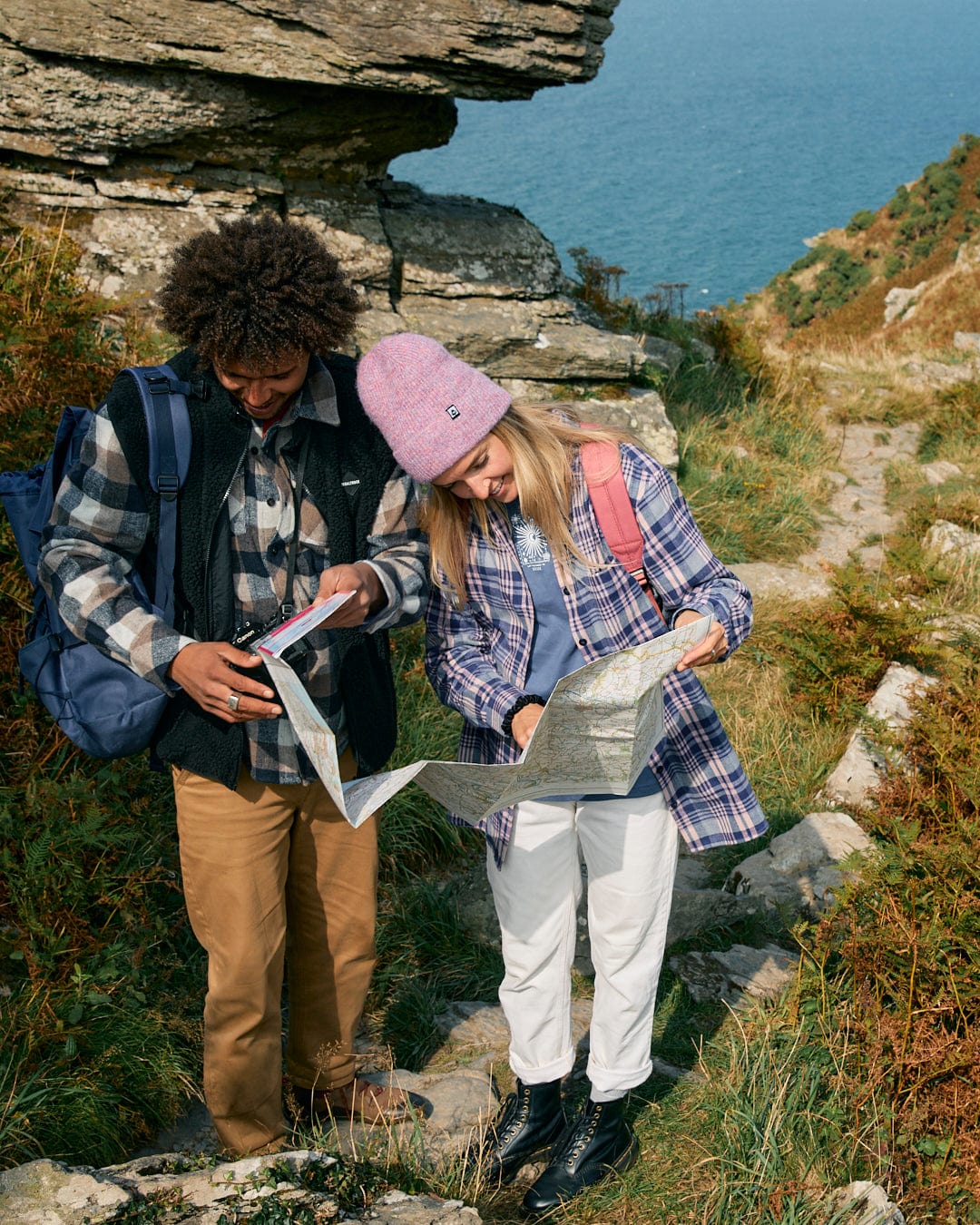 Two individuals dressed in casual attire stand on a rocky path near the ocean, examining maps while one of them is wearing the "Kizzie" women's longline shirt in purple from Saltrock.