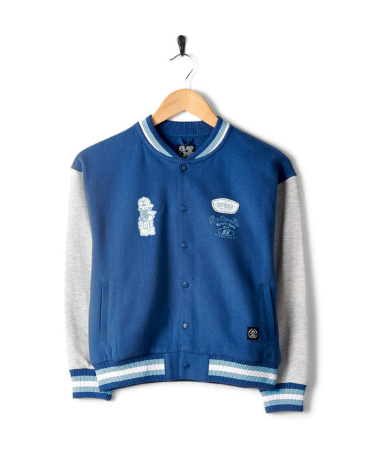 A blue and gray **Krew - Recycled Varsity Bomber Jacket - Blue** with white and blue striped cuffs and collar, featuring various patches and **Saltrock** branding, hangs on a wooden hanger on a white background.