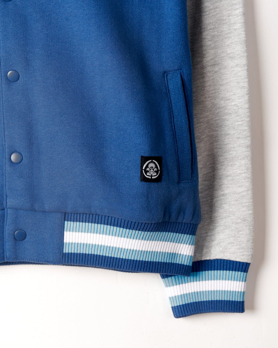 Close-up of a blue and gray Krew - Recycled Varsity Bomber Jacket - Blue with a white, blue, and turquoise striped cuff; features the Saltrock branding and a monkey head logo near the pocket.