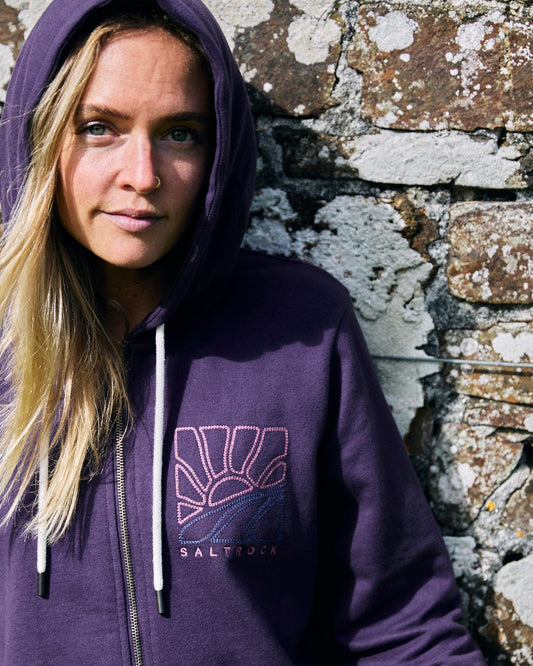 A person wearing the Saltrock Landscape Women's Zip Hoodie in purple, made from soft cotton material, stands gracefully in front of a textured stone wall.