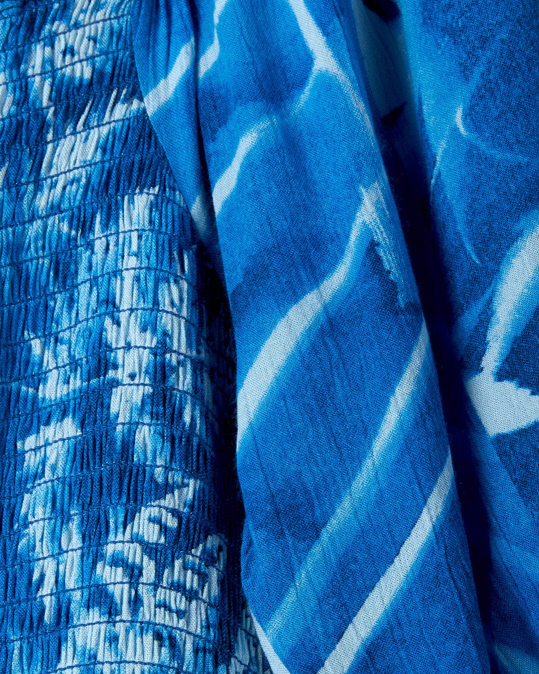 Close-up of a blue fabric with white patterns, featuring an abstract floral pattern and a mix of smooth and textured surfaces. The fabric is from the Larran Cyanotype - Midi Woven Dress - Blue by Saltrock.