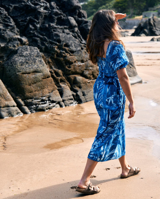 A woman in a Saltrock Larran Cyanotype - Midi Woven Dress - Blue and sandals walks along a rocky beach, shielding her eyes with her hand.