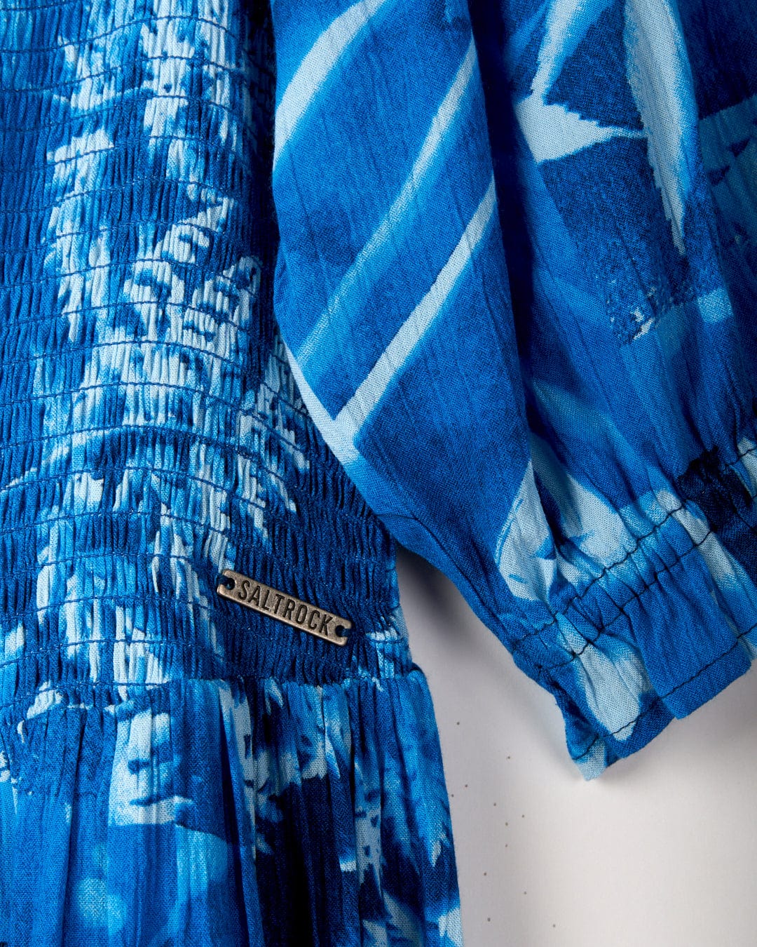 Close-up of a blue garment with a Saltrock label, featuring a smocked waist and gathering at the hem. The fabric, crafted from crinkle viscose, showcases a tie-dye pattern in various shades of blue. It is identified as the Larran Cyanotype - Midi Woven Dress - Blue from Saltrock.