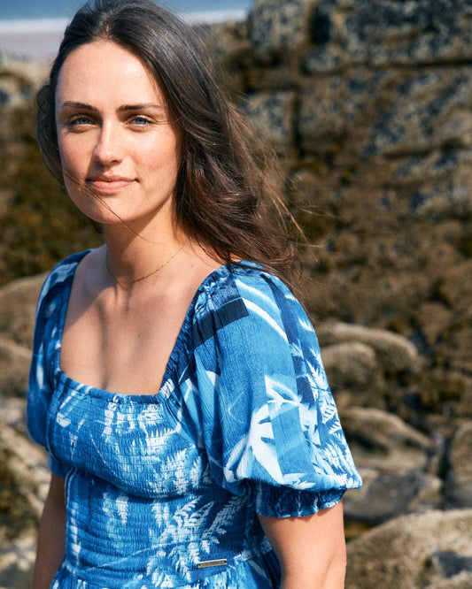 A woman with long brown hair, wearing a Saltrock Larran Cyanotype - Midi Woven Dress - Blue with elasticated hems, stands outdoors against a rocky background.