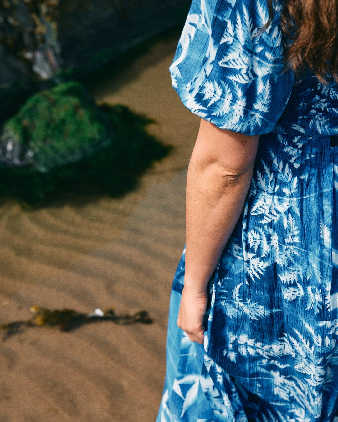 A person in a Larran Cyanotype - Midi Woven Dress - Blue from Saltrock stands next to shallow water with visible sand patterns and green mossy rocks.