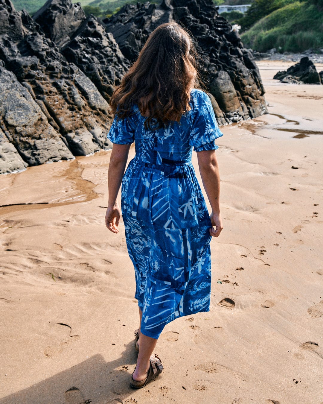 A person with long hair, wearing a Larran Cyanotype - Midi Woven Dress - Blue from Saltrock, walks on a sandy beach toward rocky formations.