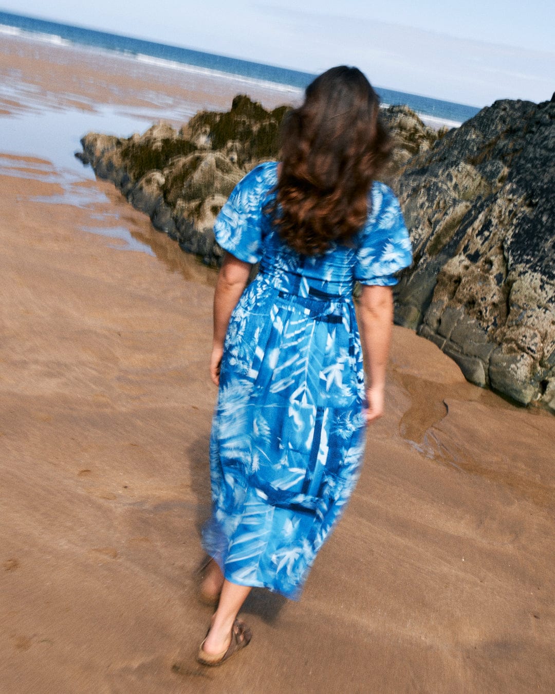 A person with long hair, wearing a Larran Cyanotype - Midi Woven Dress - Blue from Saltrock, walks barefoot on a sandy beach near rocky formations.