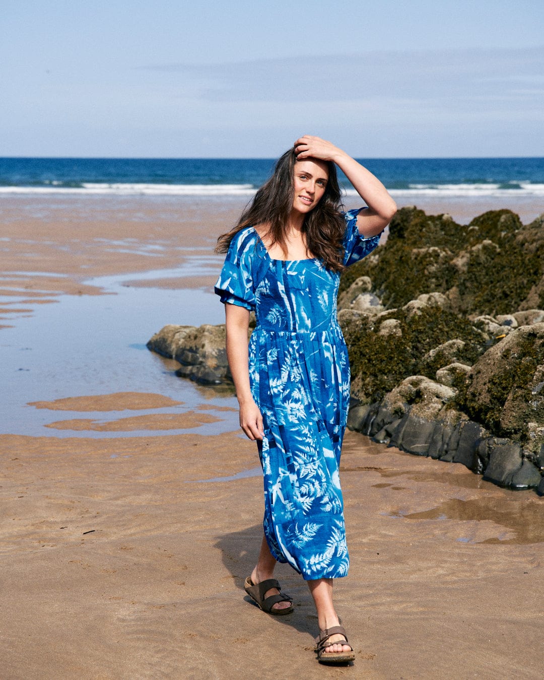 A woman in a Larran Cyanotype - Midi Woven Dress - Blue by Saltrock stands on a sandy beach near the ocean, touching her hair with one hand, with rocks visible in the background.