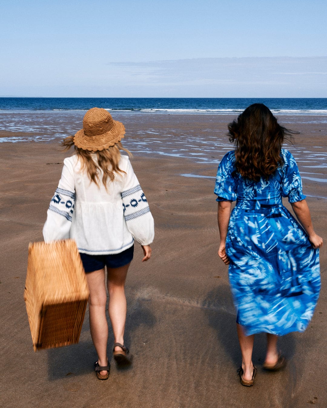 Two women walk on a sandy beach toward the ocean. One carries a wicker basket, and both wear casual beach attire, including the Saltrock Larran Cyanotype - Midi Woven Dress - Blue.