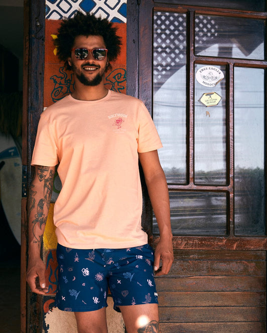 A man with curly hair and sunglasses stands outside a wooden door wearing a light orange T-shirt and Saltrock's Last Stop - Mens Swimshorts - Blue featuring an elasticated waist.