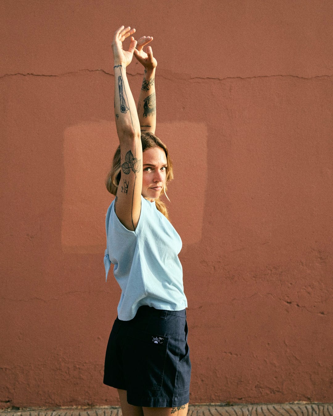 A person with tattoos on their arms stretches upward against a textured red wall, wearing a light blue 100% cotton sleeveless shirt and Liesl - Womens Chino Shorts - Blue from Saltrock.