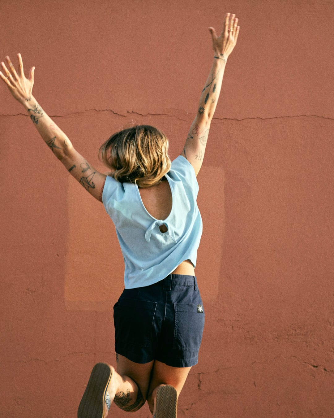 Person in a blue top and Saltrock Liesl - Womens Chino Shorts - Blue, made of machine washable cotton twill, jumping with arms raised against a pinkish-brown wall. Tattoos are visible on their arms and legs.