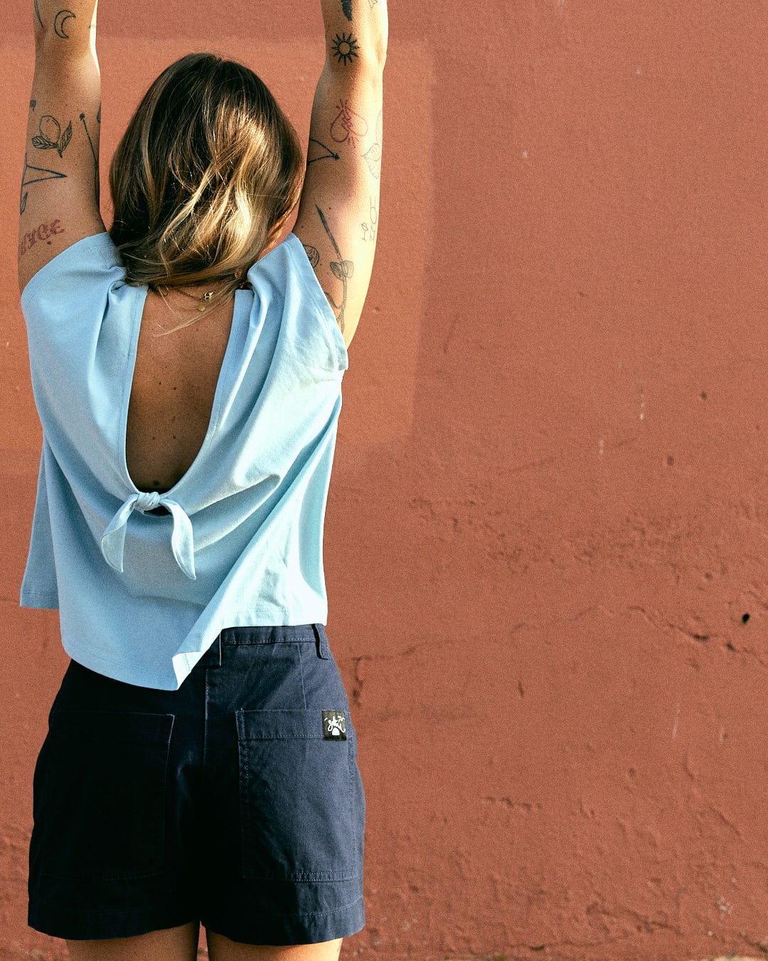 Person with arm tattoos stretches in front of a pink wall, wearing a light blue top with an open back and high-waisted Liesl - Womens Chino Shorts - Blue made from cotton twill by Saltrock. The ensemble is machine washable.