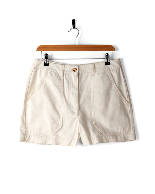 A pair of Liesl - Womens Chino Short - White is hanging on a wooden hanger, featuring side pockets and a front button closure. These stylish Saltrock shorts combine comfort and practicality.