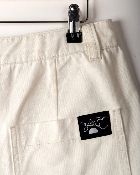 Close-up of an off-white cotton twill garment with a black "Saltrock" label sewn onto a pocket. The Liesl - Womens Chino Short - White by Saltrock are high-waisted shorts hanging on a metal clip.